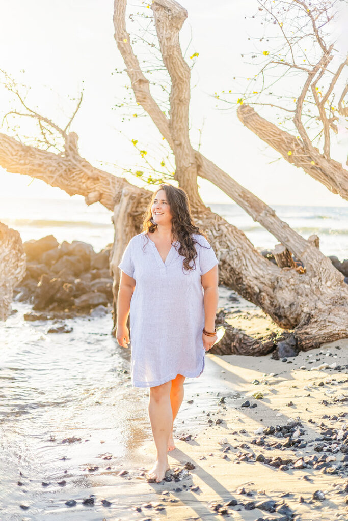 woman in a blue dress walking on th sandy beach with driftwood during sunset 