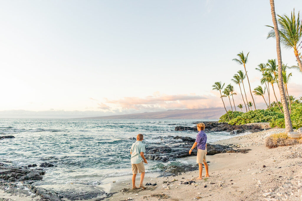 kids throwing rocks into the ocean standing on a sandy beach with lava rocks and palm trees.